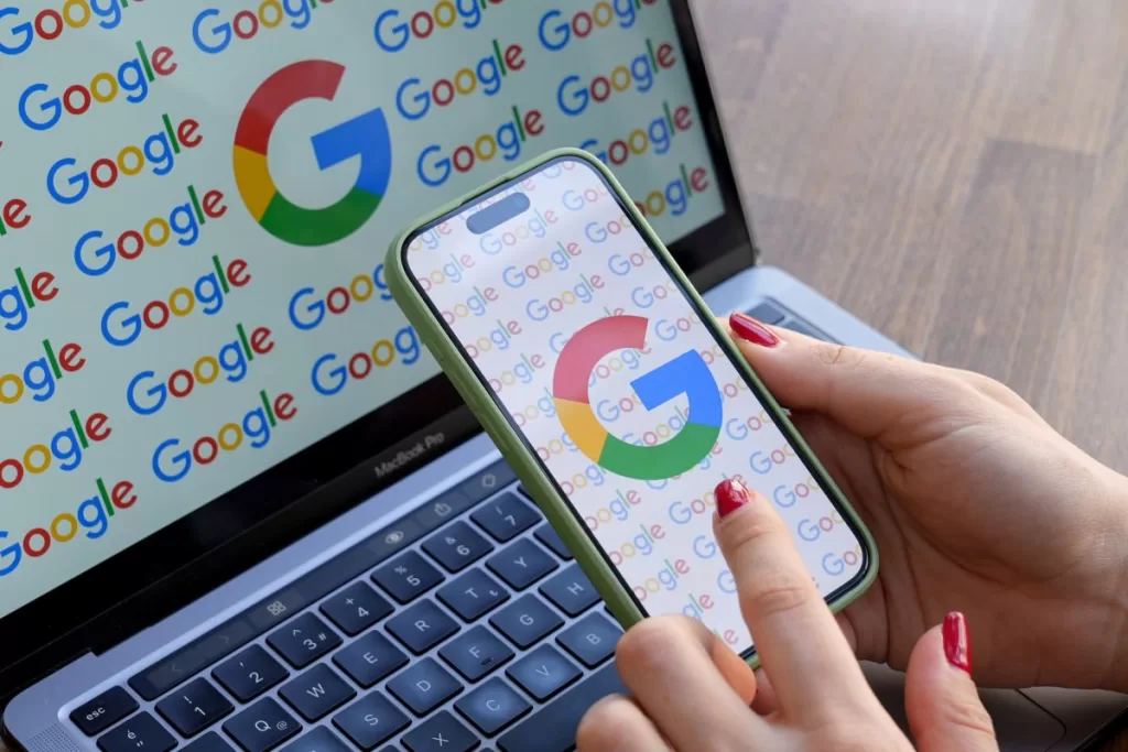 A woman looks at a mobile phone displaying the logo of Google in front of a laptop screen displaying the logo of Google.