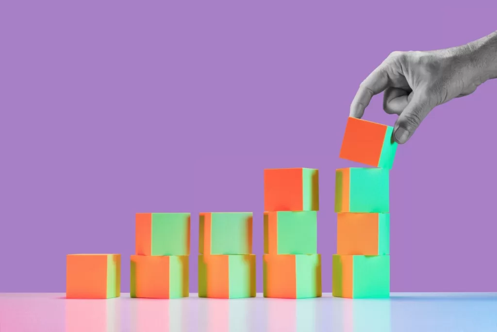 Man's hand in black and white placing block on largest stack of 5 growing stacks of yellow and red tinted blocks